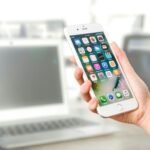 Close-up view of a smartphone displaying apps, held by a hand, with a blurred laptop in the background.